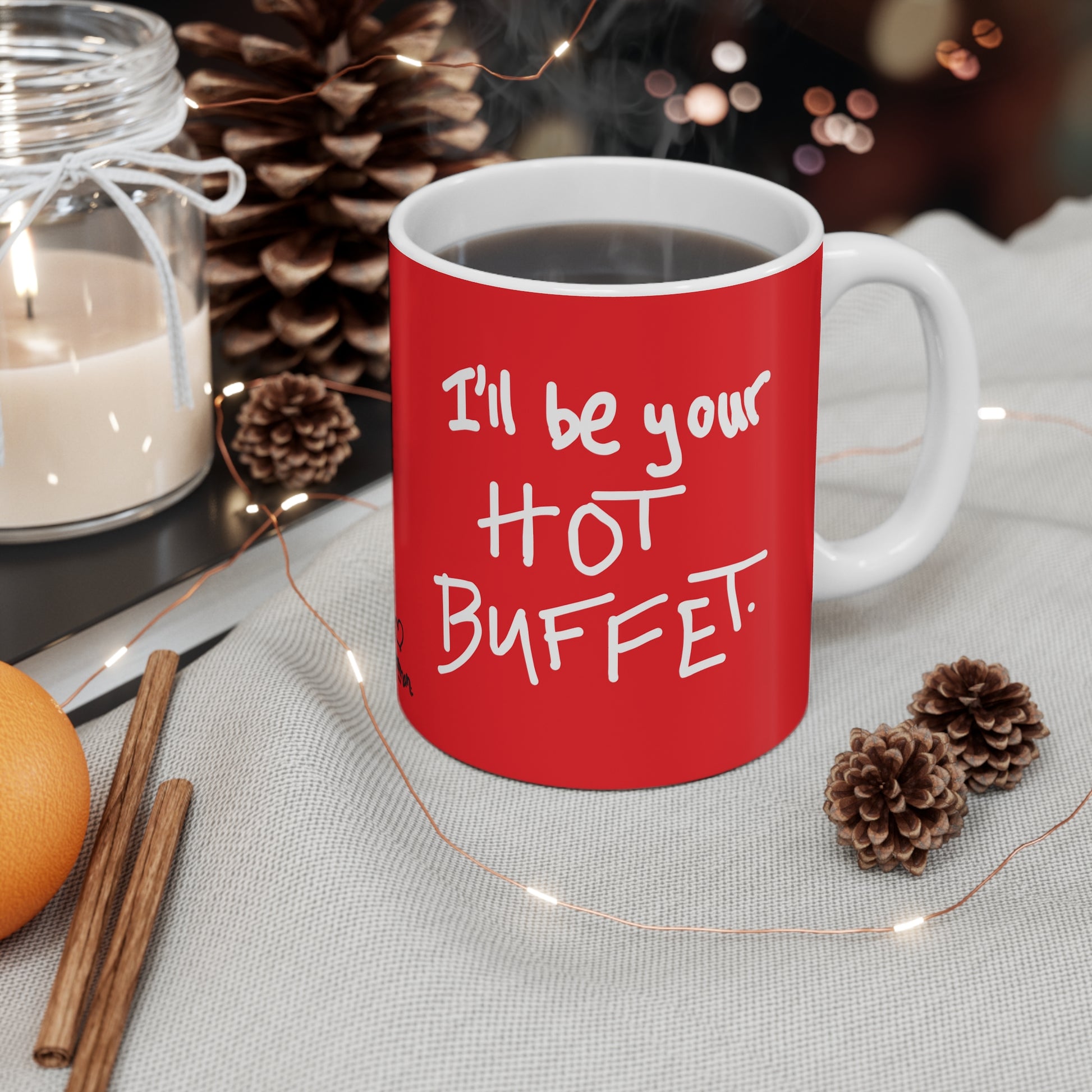 Red mug with 'I'll be your hot buffet' text on a table with decorative items.