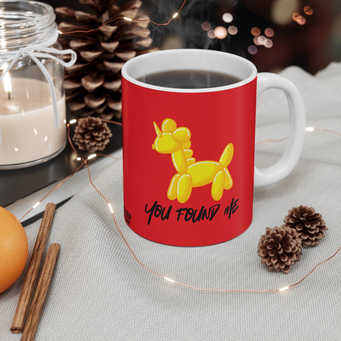 Red mug with yellow balloon unicorn design and text 'You Found Me' on a table with decorative items.